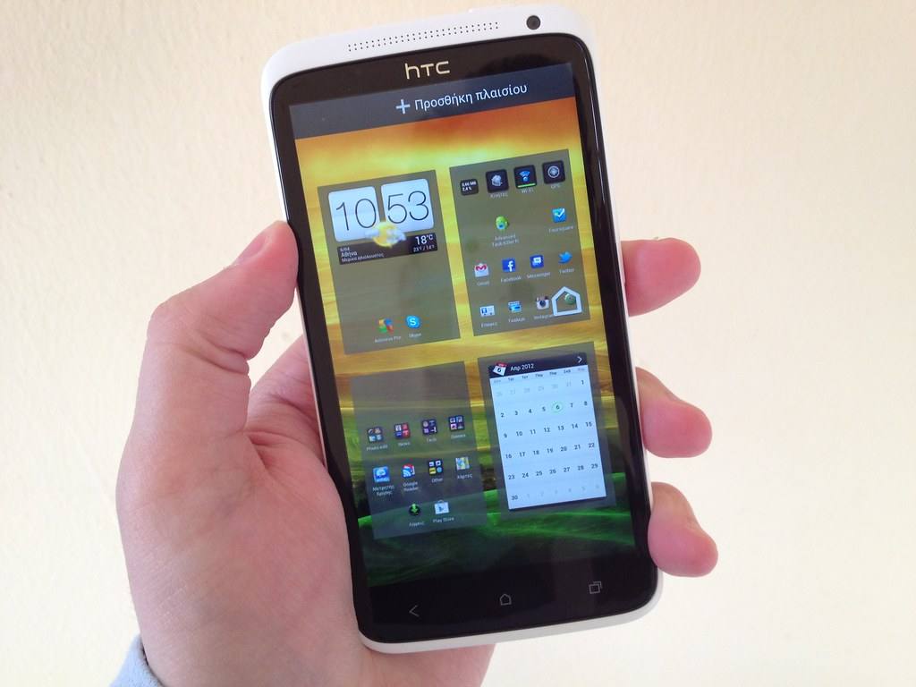 Bílý telefon HTC One X v ruce, na displeji domovská obrazovka s hodinami a kalendářem