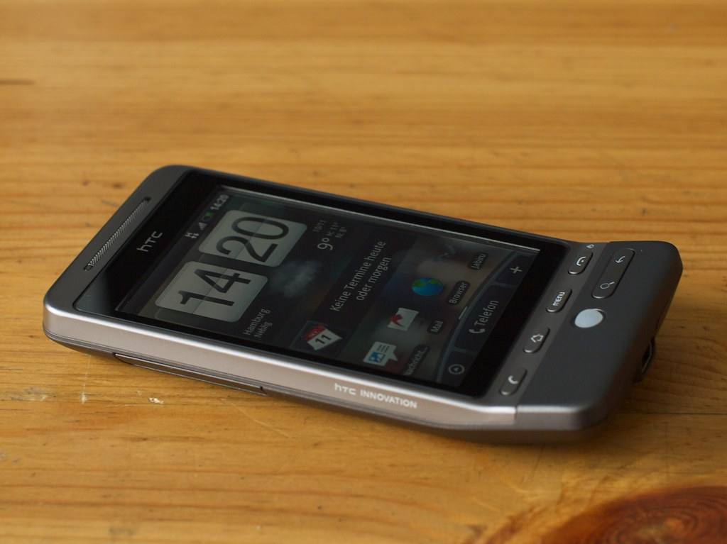 Telefon HTC Hero ležící na dřevěném stole, na displeji hodiny nadstavby Sense