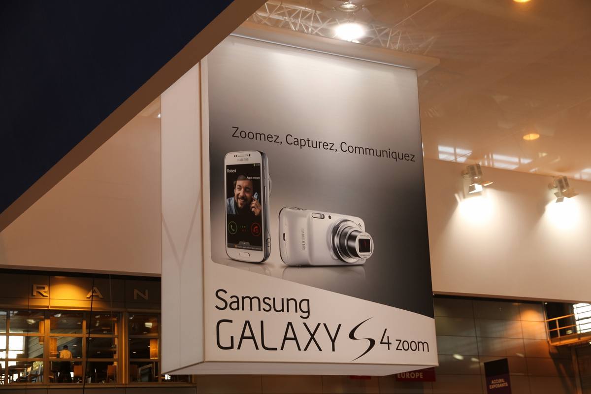 Reklamní poutač na Galaxy S4 Zoom, na kterém je telefon zezadu s vysunutým tubusem objektivu