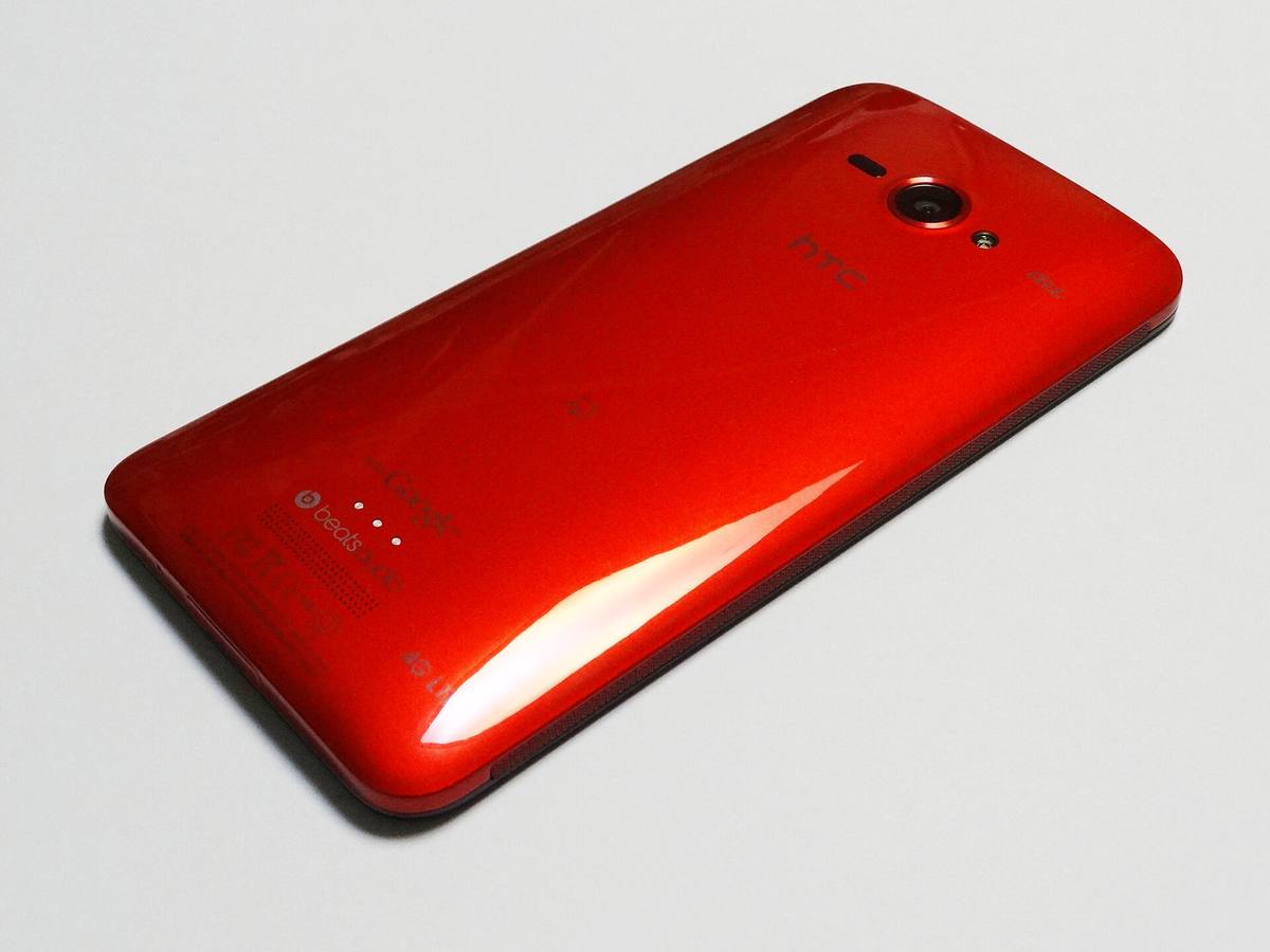 Záda červeného telefonu HTC J butterfly ležícího na bílém podkladu, vidět je fotoaparát a nápis HTC