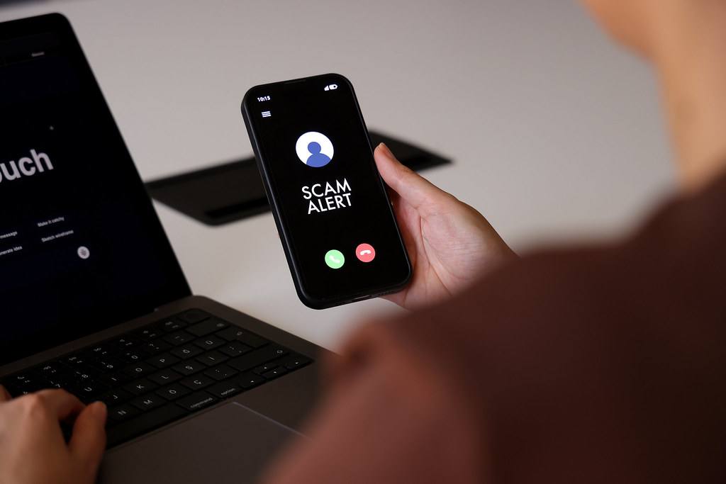 Ruka drží telefon, na displeji je příchozí hovor s nápisem Scam Alert