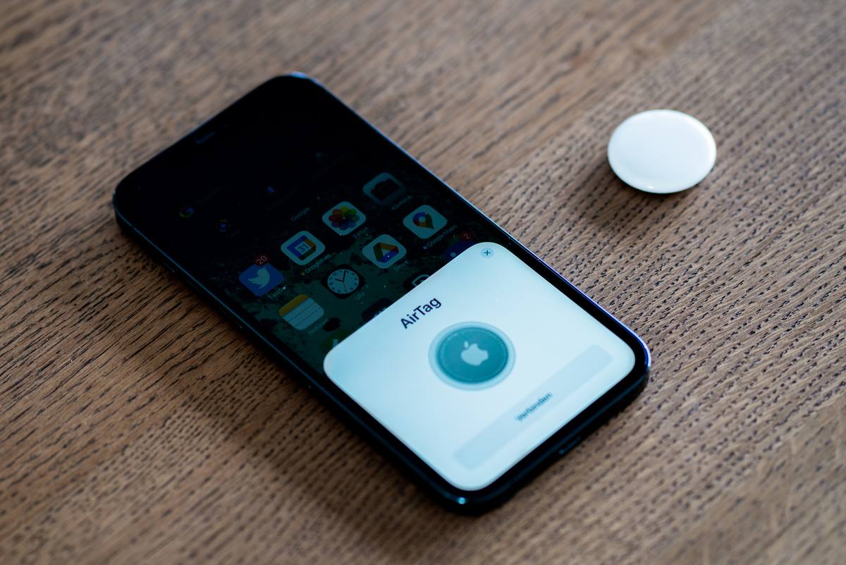 Bílý kulatý přívěsek AirTag leží vedle iPhonu, na jehož displeji běží nastavení