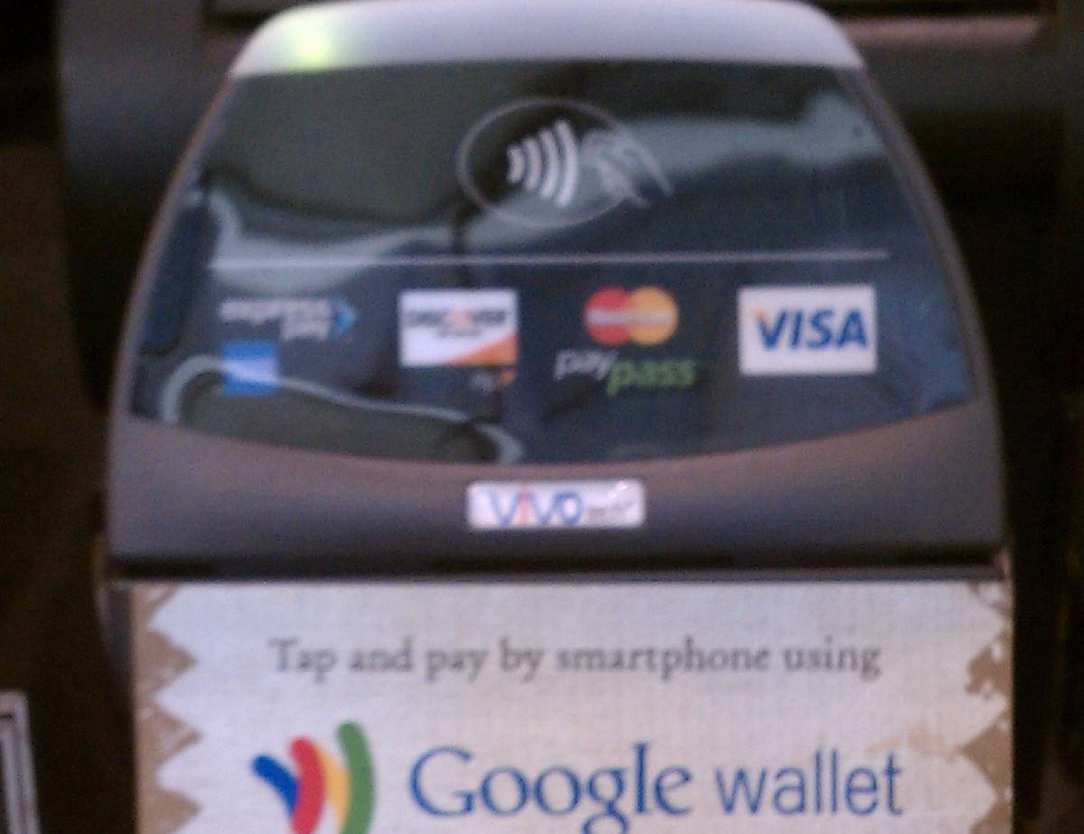 Bezkontaktní platební terminál s cedulkou vyzývající k placení telefonem přes Google Wallet