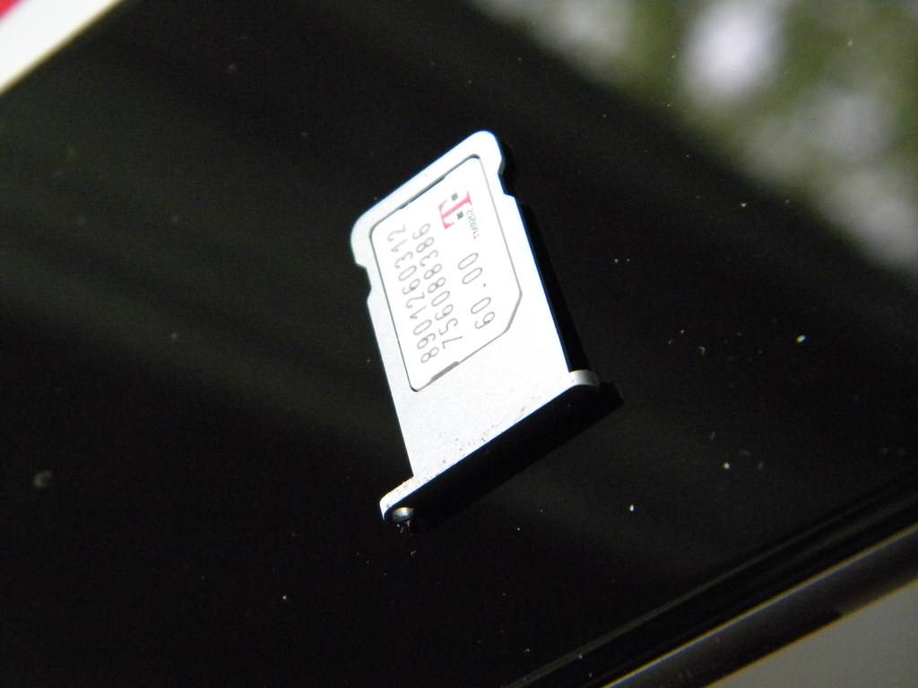 Stříbrný šuplík na SIM kartu z iPhonu s vloženou nanoSIM kartou s logem T-Mobile, položený na tmavém skle telefonu