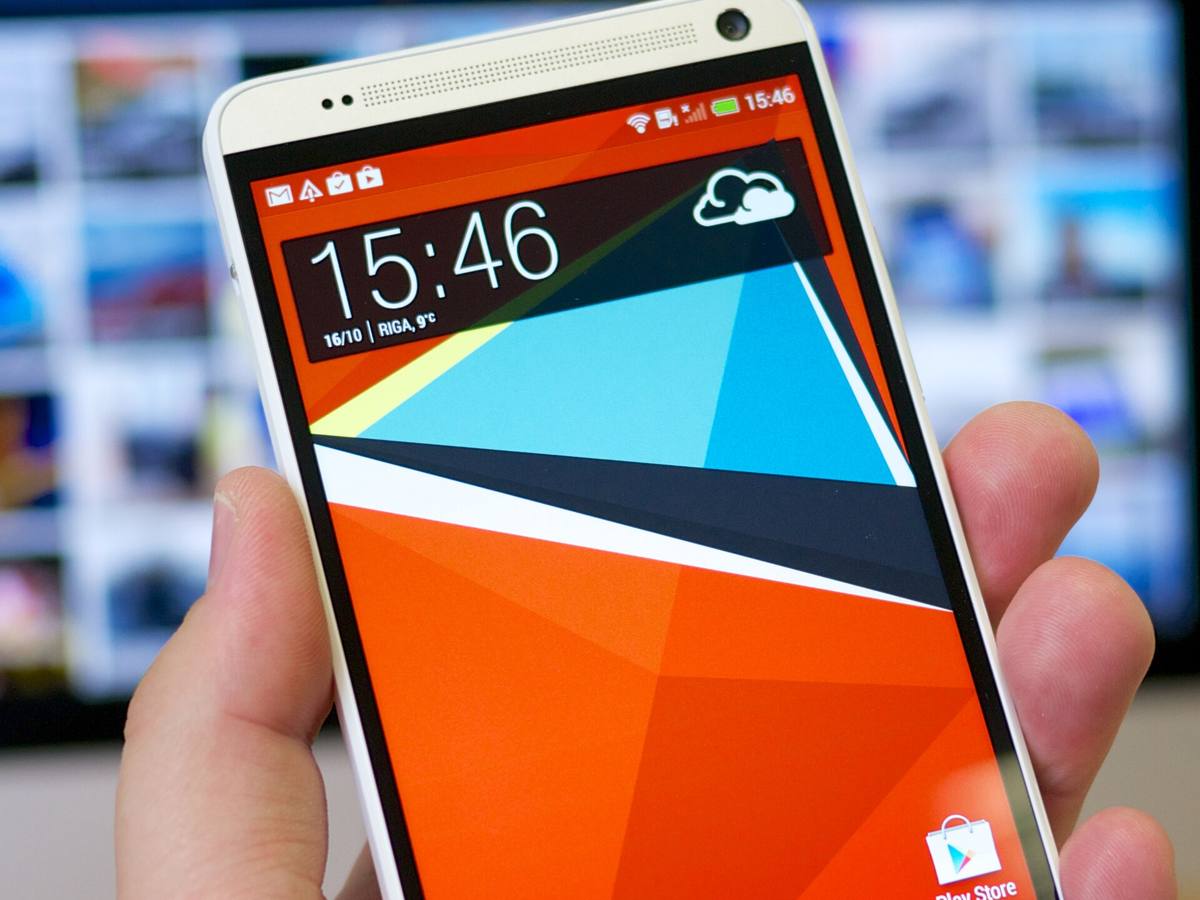 Bílý telefon HTC One max držený v ruce, na displeji domovská obrazovka s hodinami