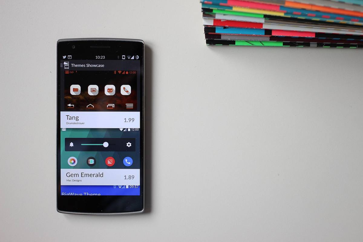 Telefon OnePlus One ležící na světlém stole vedle stohu barevných časopisů, na displeji obchod s motivy