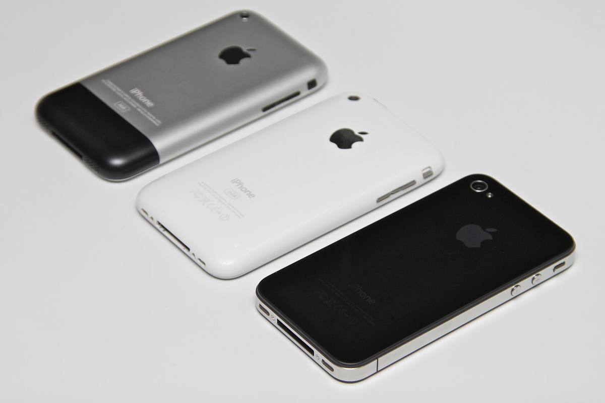 Záda tří iPhonů vedle sebe na bílém pozadí, hliníkový původní model, bílý 3G a černý iPhone 4